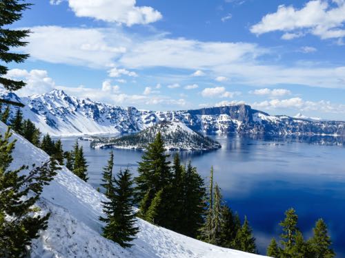 Snow covered island in a blue lake surrounded by mountains with snow on an off-the-beaten-path national park trip to Crater Lake National Park.