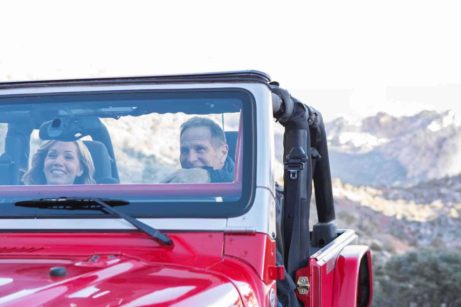 Man and woman sitting in a red jeep with mountains in the background