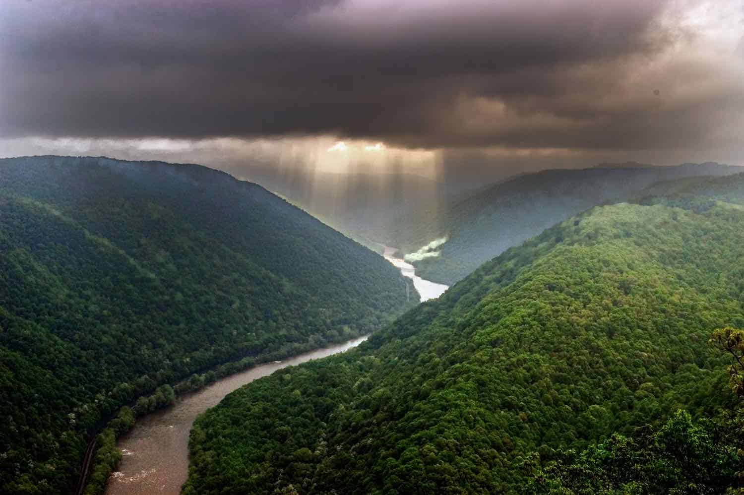 Light shafts stream through a storm cloud illuminating a river that flows between green mountains at New River Gorge National Park.
