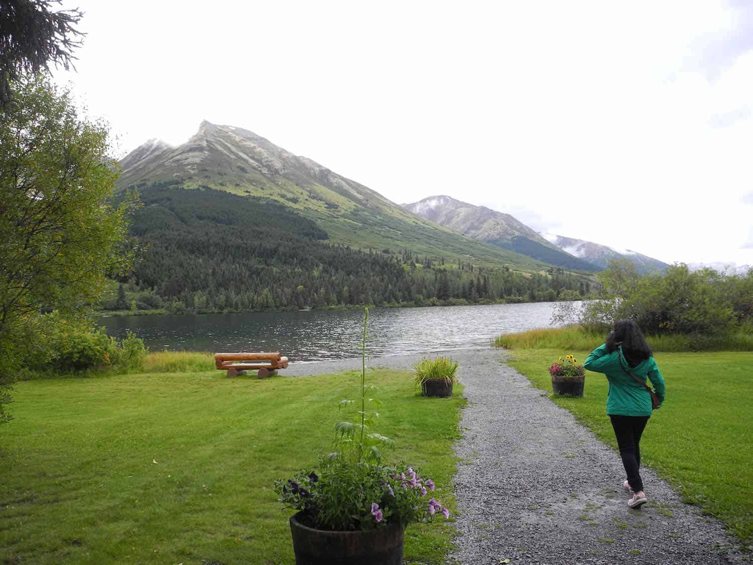 woman walking down a path to a lake surrounded by green lawn and mountains