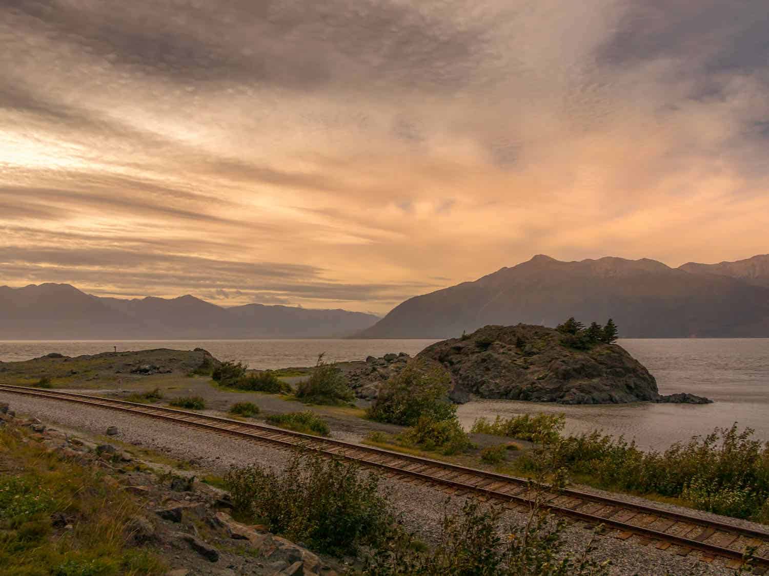 train tracks traveling next to Turnagain Arm on the Seward Highway in Alaska. Mountains in the distance and a cloudy sky with streaks of pale light.