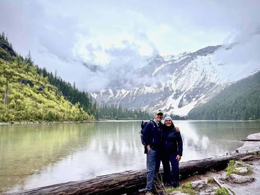 Couple standing in front of Avalanche Lake with snow-covered mountains behind them.