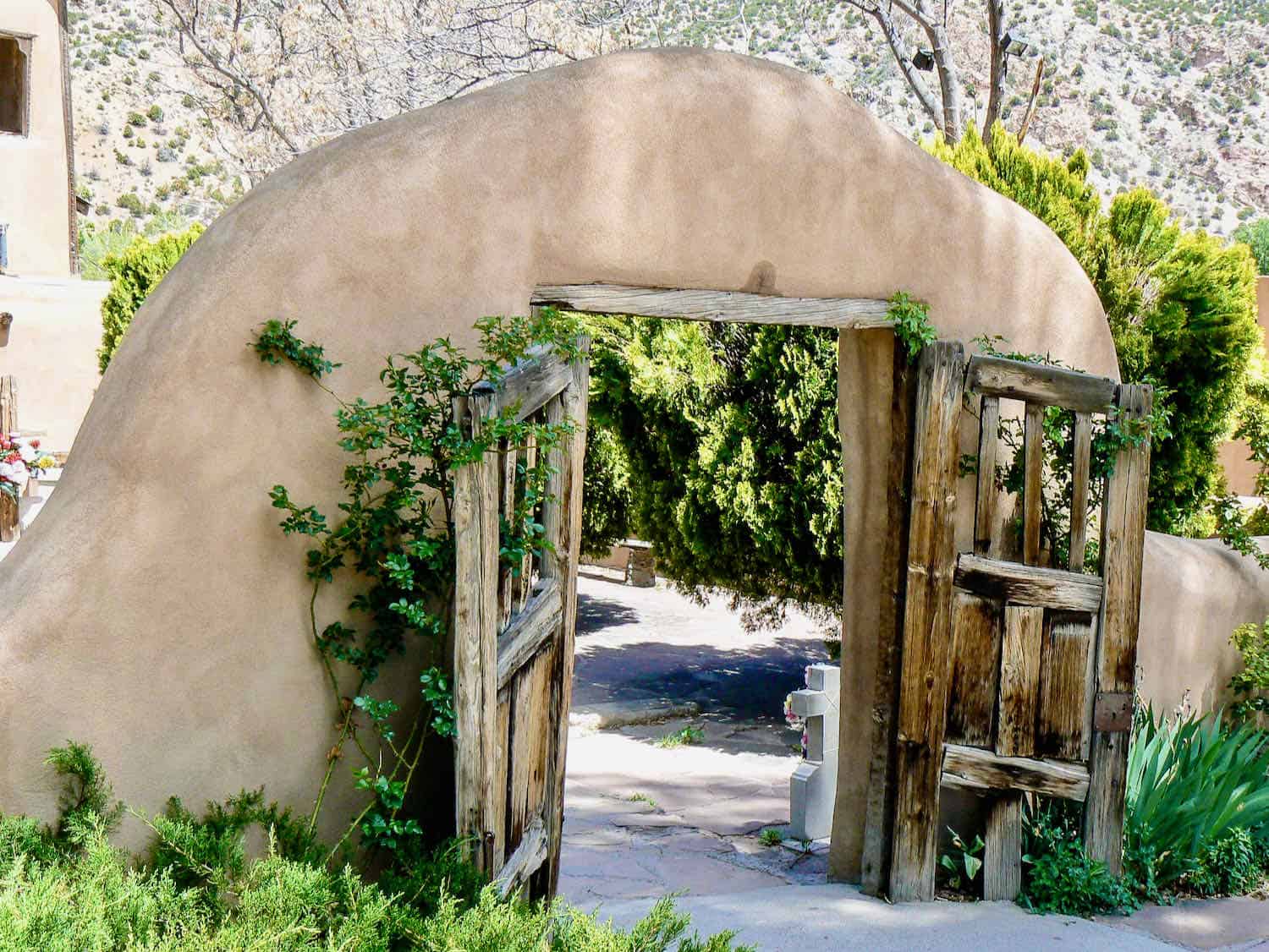 Wooden gates in an adobe arched entry way surrounded by greenery.
