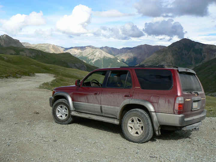 Red SUV covered in dirt on a four-wheel-drive-trail with mountains in the distance