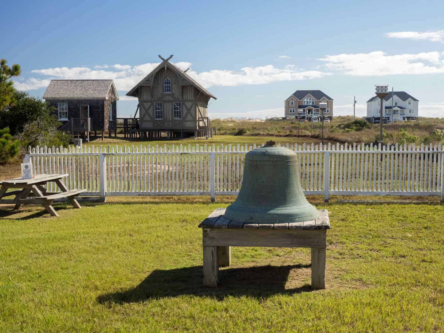 Big bell sitting in front of a white picket fence with four buildings behind the fence.