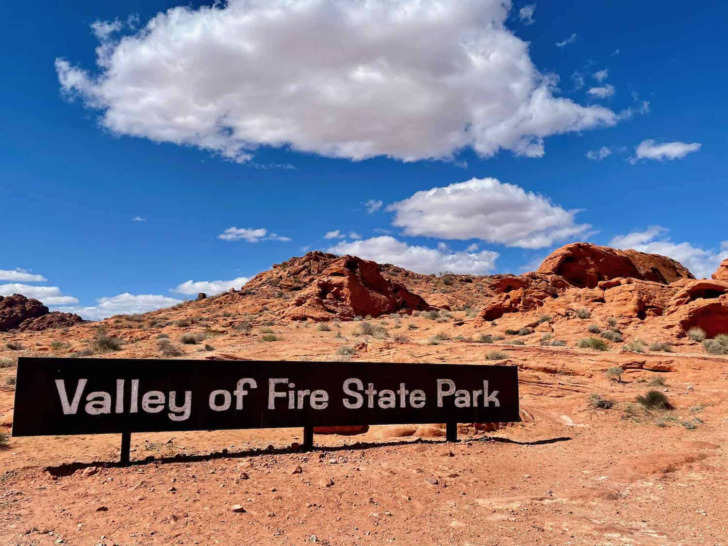 Sign that says Valley of Fire State Park with red rocks surrounding it.