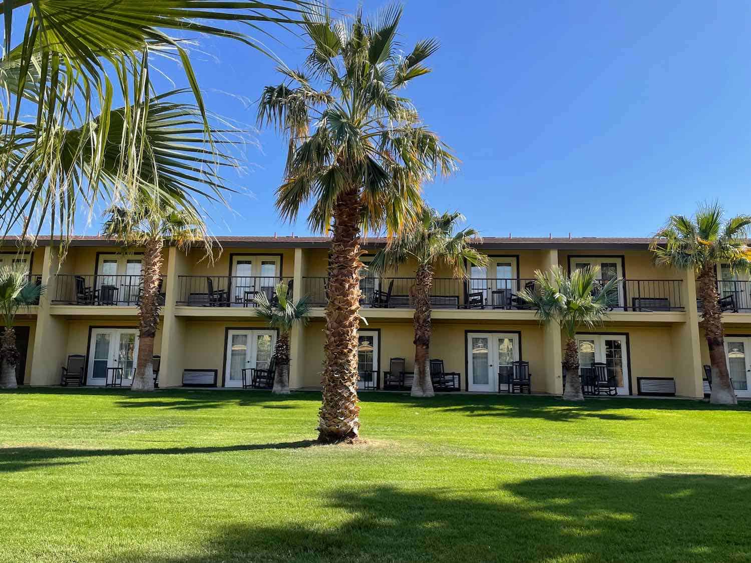 Low-slung two-story yellow buildings surrounded by green grass and palm trees at The Ranch at Death Valley.