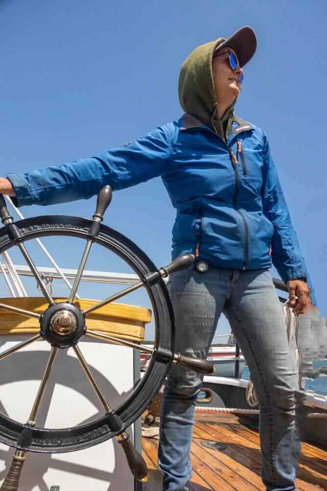 Woman in a blue jacket and wearing a hat stands at the wheel of a Maine windjammer schooner.
