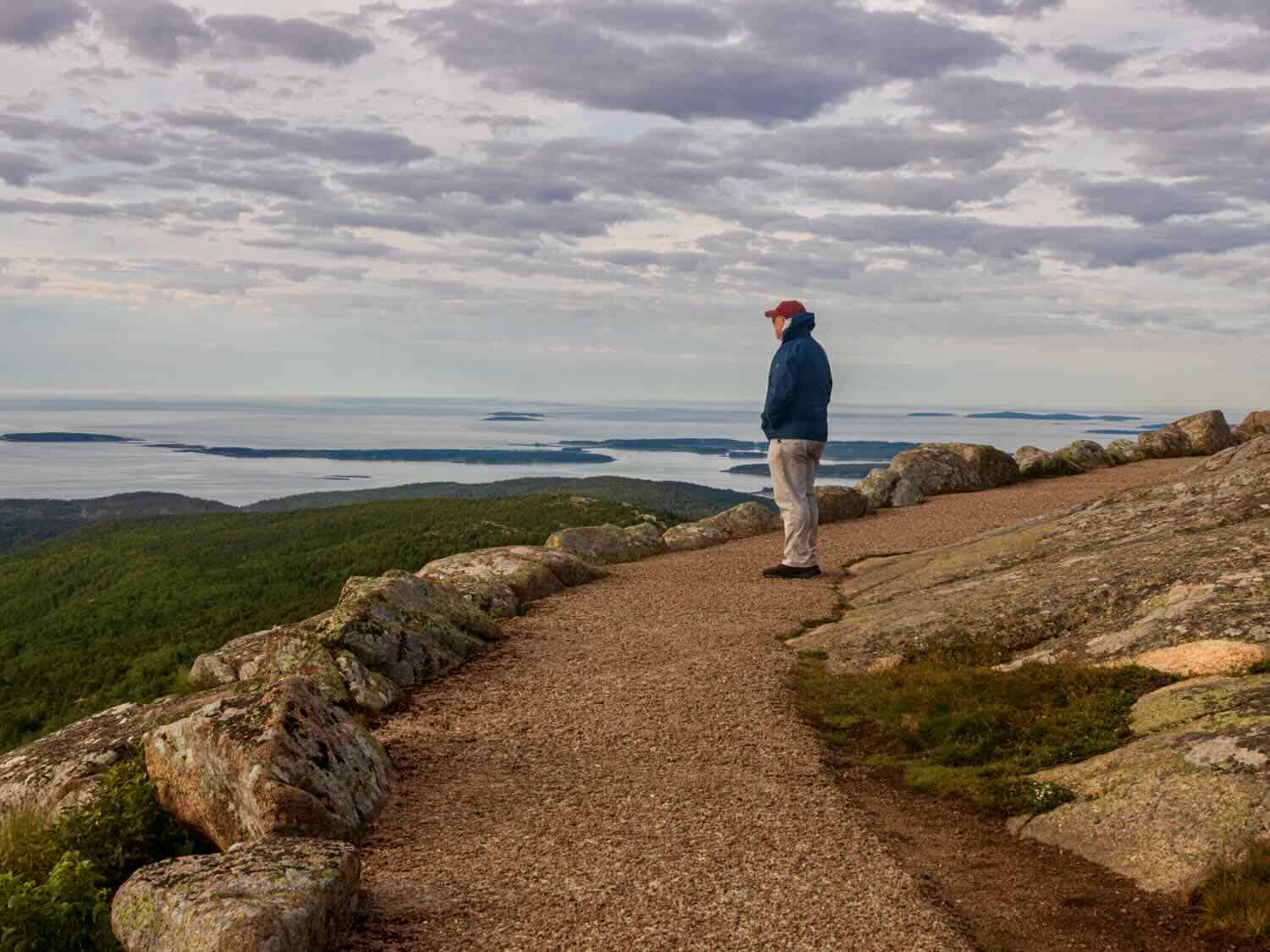Man stands on a trail taking in the view of ocean and islands at Cadillac Mountain in Acadia National Park.