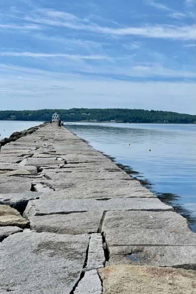 A wide rock pier surrounded by water with Rockland Breakfront Lighthouse sitting at the end of the pier.