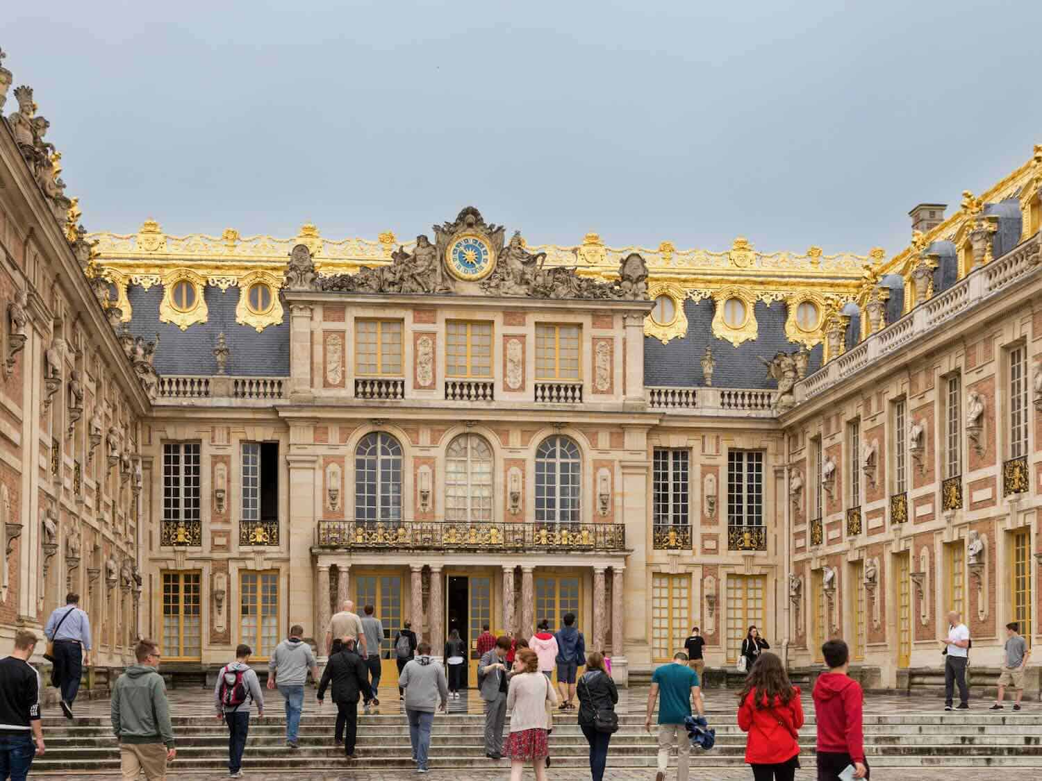 A crowd walks up steps toward the ornate Palace of Versailles with a slate roof trimmed in gold.